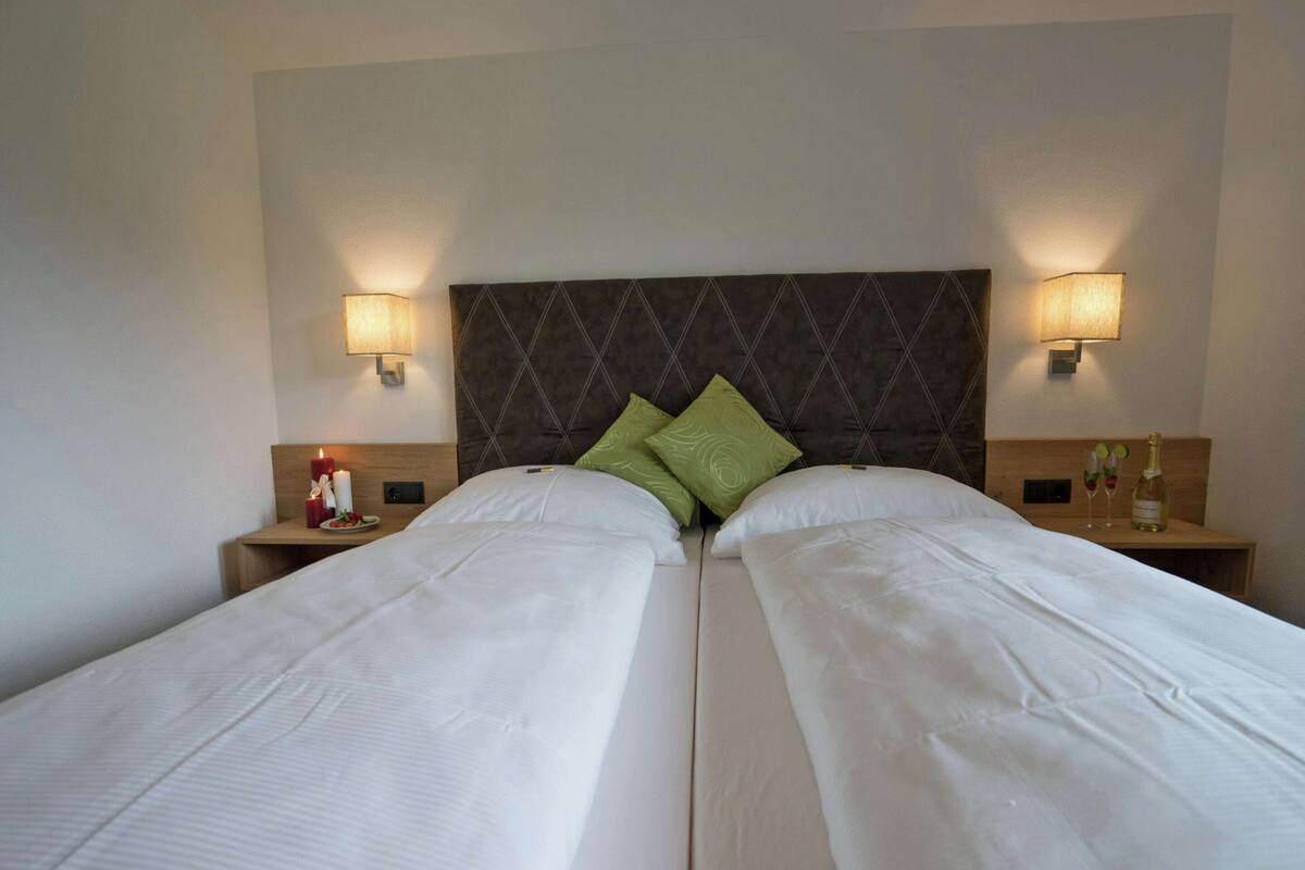 A double bed with white linens, two green pillows, and a dark headboard. There are two bedside tables with lamps and a bottle of champagne.