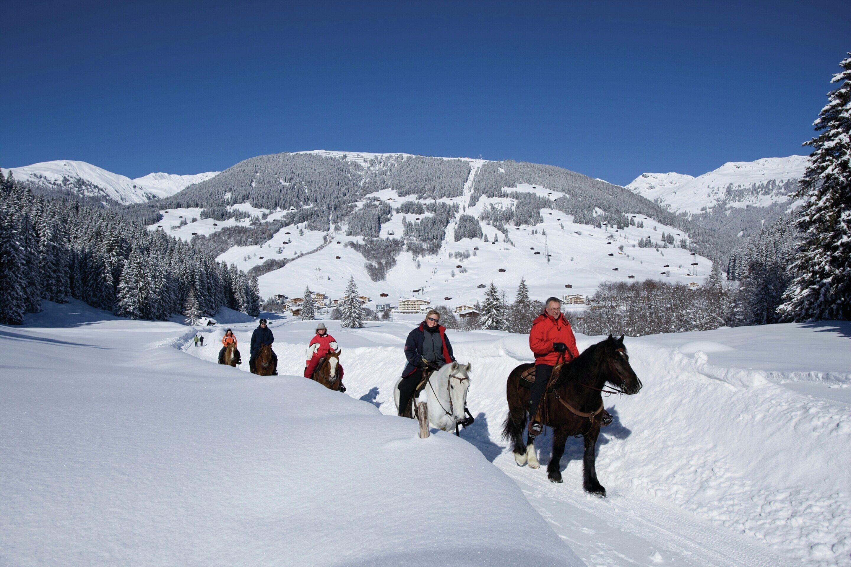 A group of people riding horses on a snow-covered path with a mountainous backdrop.
