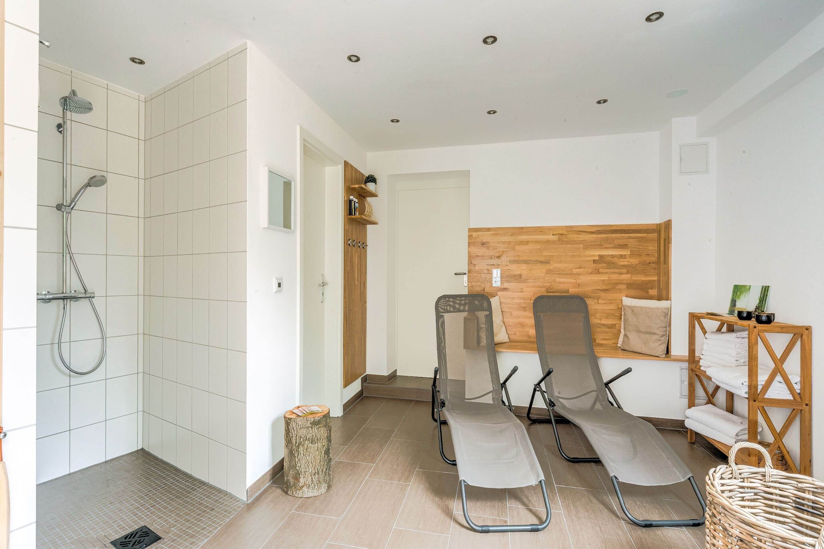 A modern bathroom with a shower, two reclining chairs, a wooden wall panel, and a shelf with towels.
