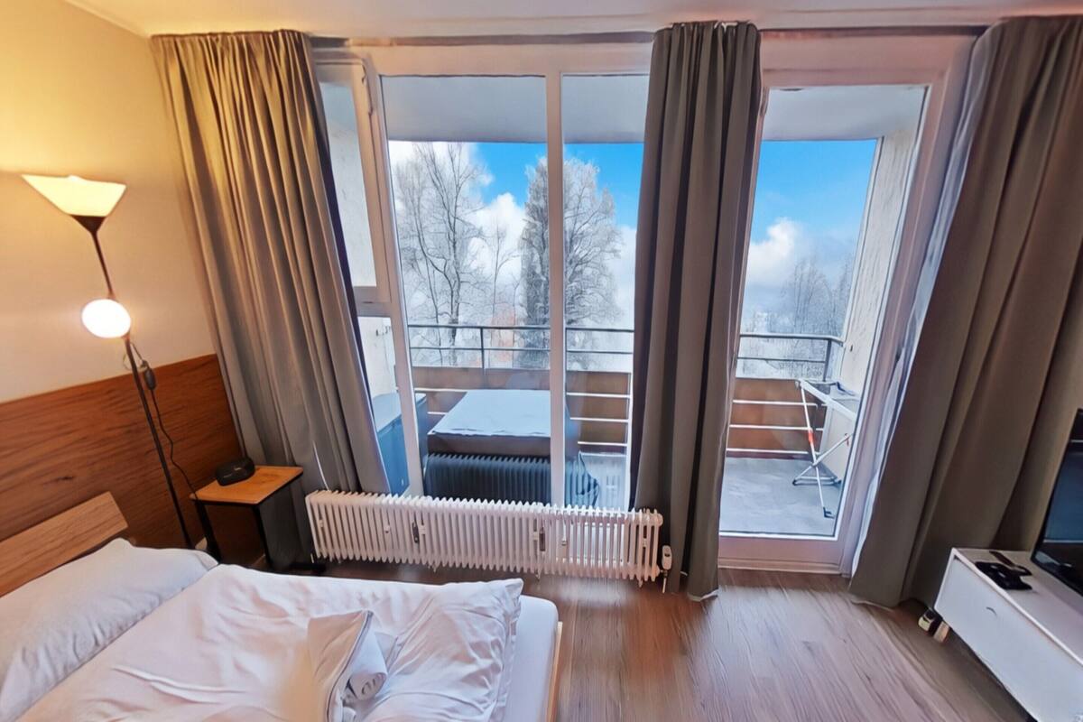 Habitación de hotel con cama, escritorio con lámpara, radiador, televisor y balcón con vistas a árboles y cielo despejado.