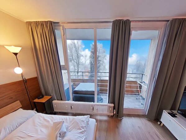 Camera d'albergo con un letto, una scrivania con una lampada, un termosifone, una televisione e un balcone affacciato su alberi e un cielo sereno.