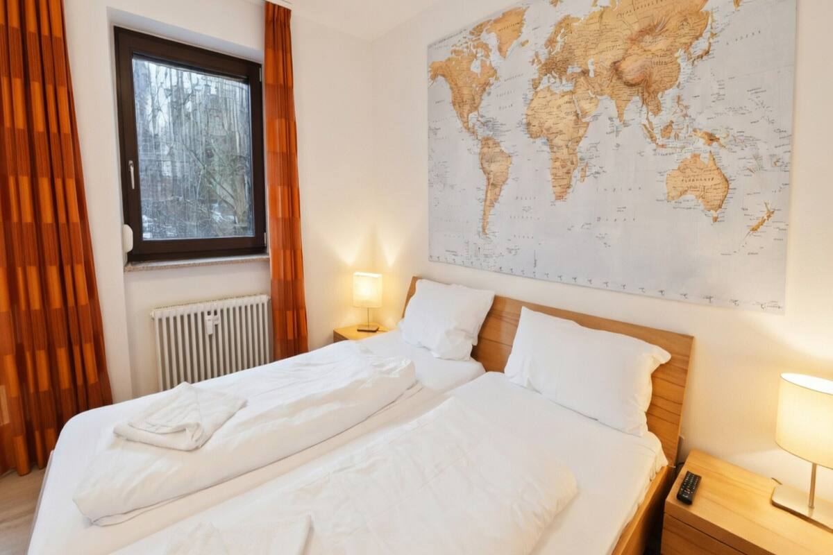 Una habitación de hotel con un gran mapa del mundo en la pared, una cama con ropa blanca y una ventana con cortinas, además de una lámpara de noche.