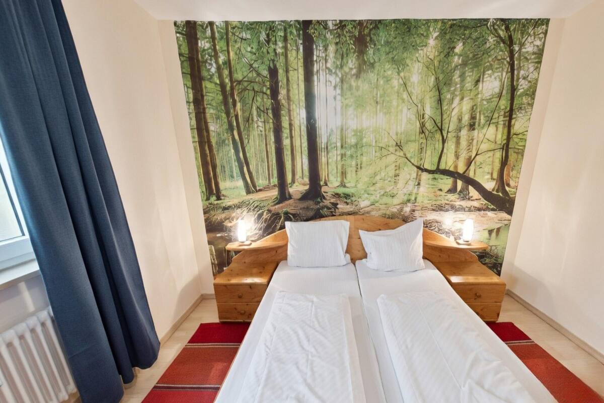 Un dormitorio con un mural temático de bosque, una cama con ropa blanca, dos mesitas de noche con velas y un ventanal con cortinas.