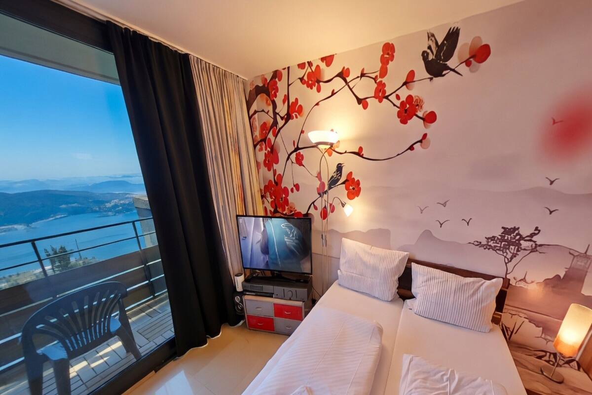 Habitación de hotel con cama, televisor, balcón con una silla y un mural en la pared.