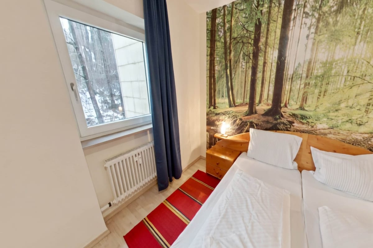 Een slaapkamer met een bed, een raam, een radiator, een rood-geel gestreept tapijt en een wandtapijt met een bosmotief.