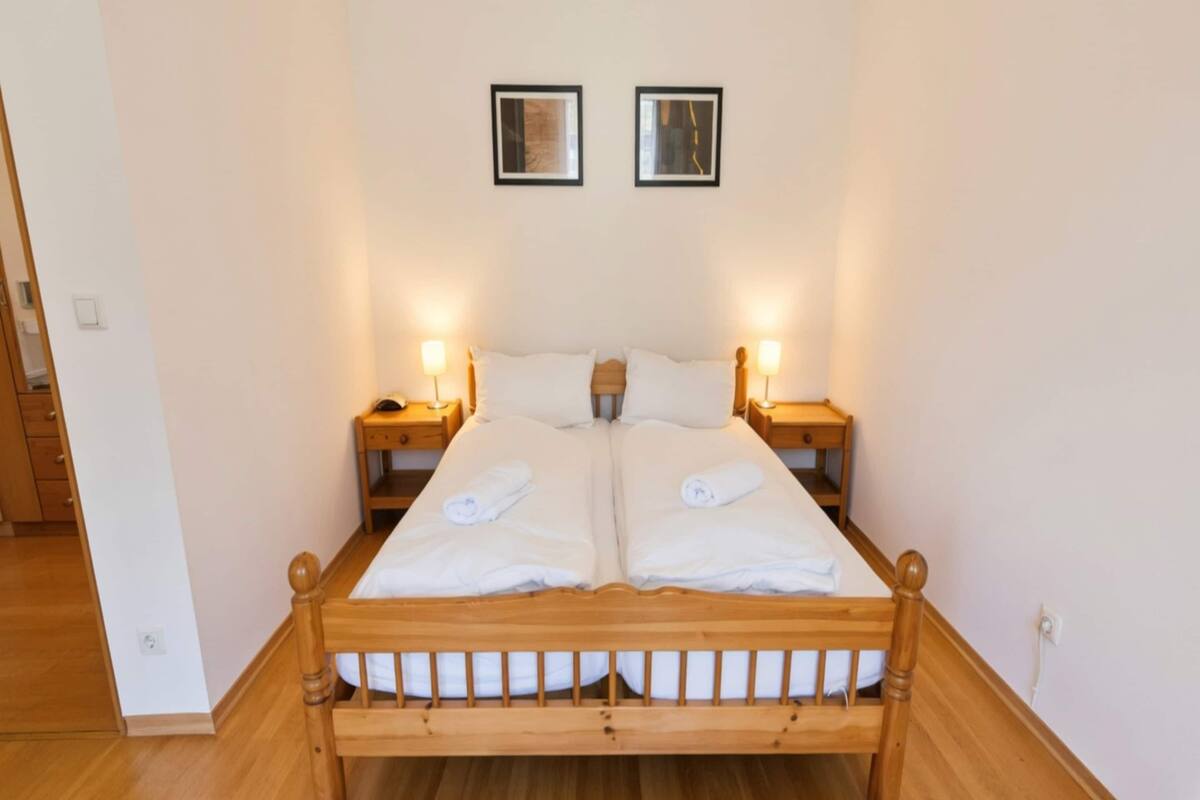 Una cama doble con cabecera de madera, dos mesitas de noche con lámparas, y dos cuadros enmarcados en la pared.