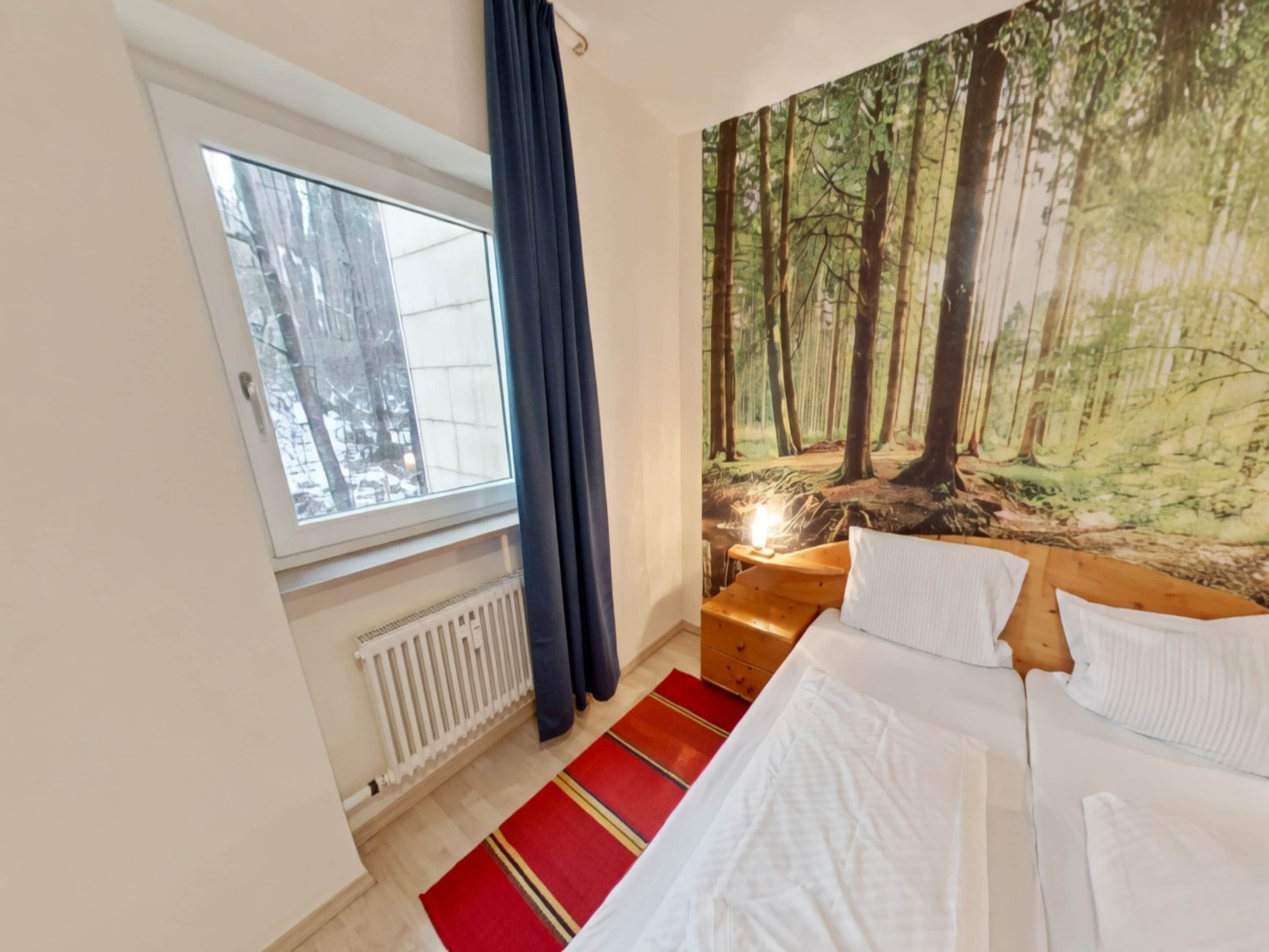 Ett sovrum med en säng, ett fönster, ett element, en röd- och gulrandig matta och en väggbonad med skogsmotiv.
