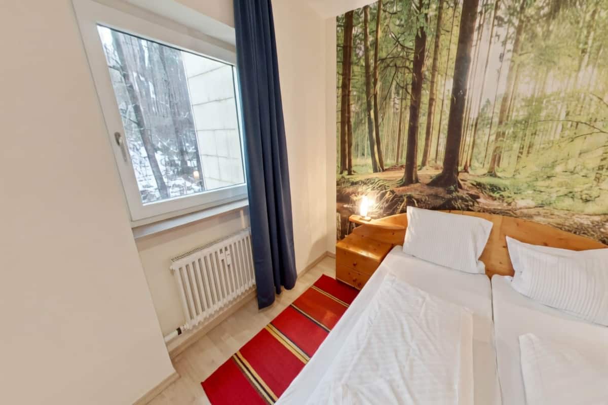 Ett sovrum med en säng, ett fönster, ett element, en röd- och gulrandig matta och en väggbonad med skogsmotiv.