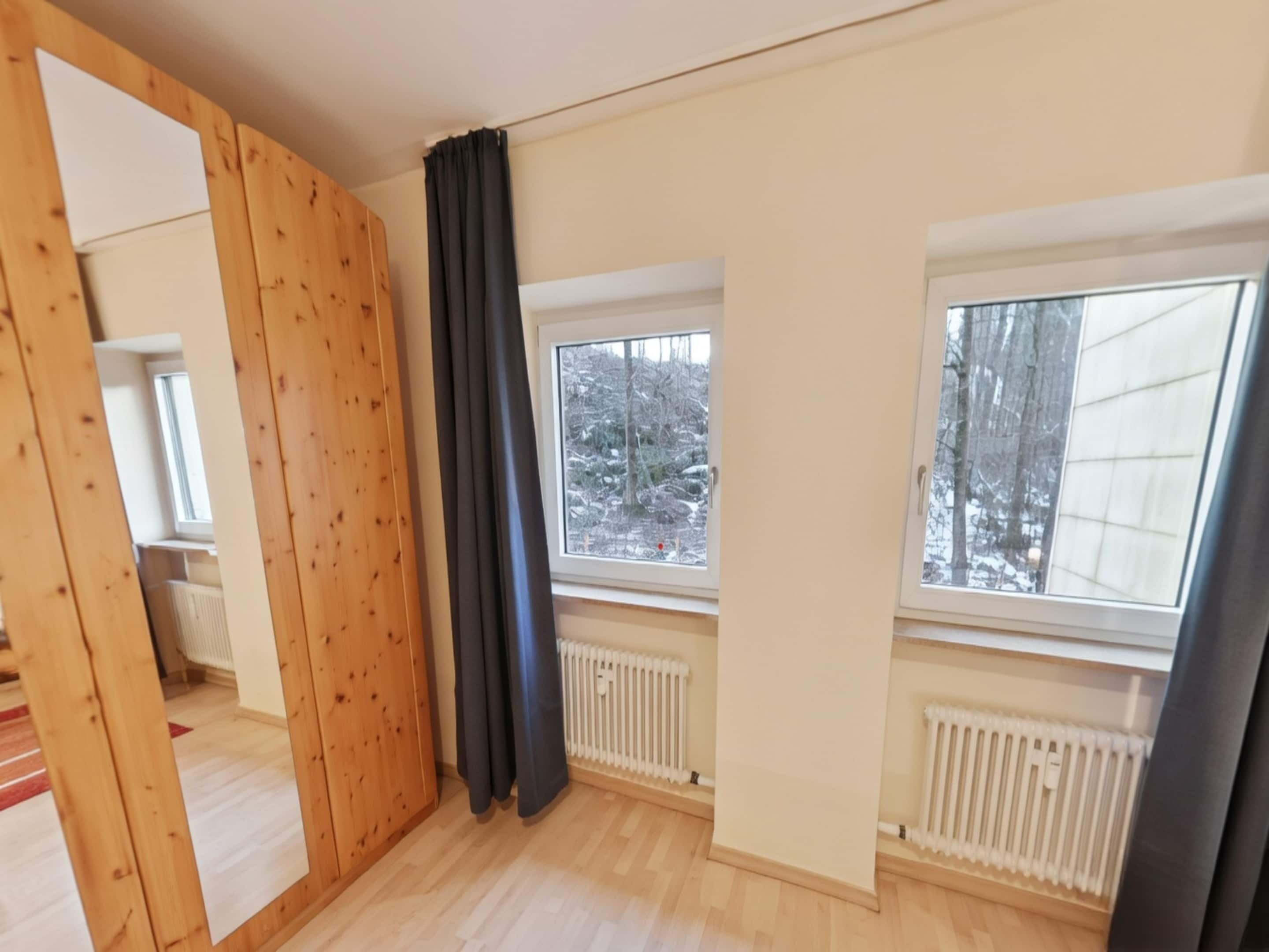 Une pièce aux murs en bois, avec deux fenêtres donnant sur des arbres couverts de neige et un radiateur.