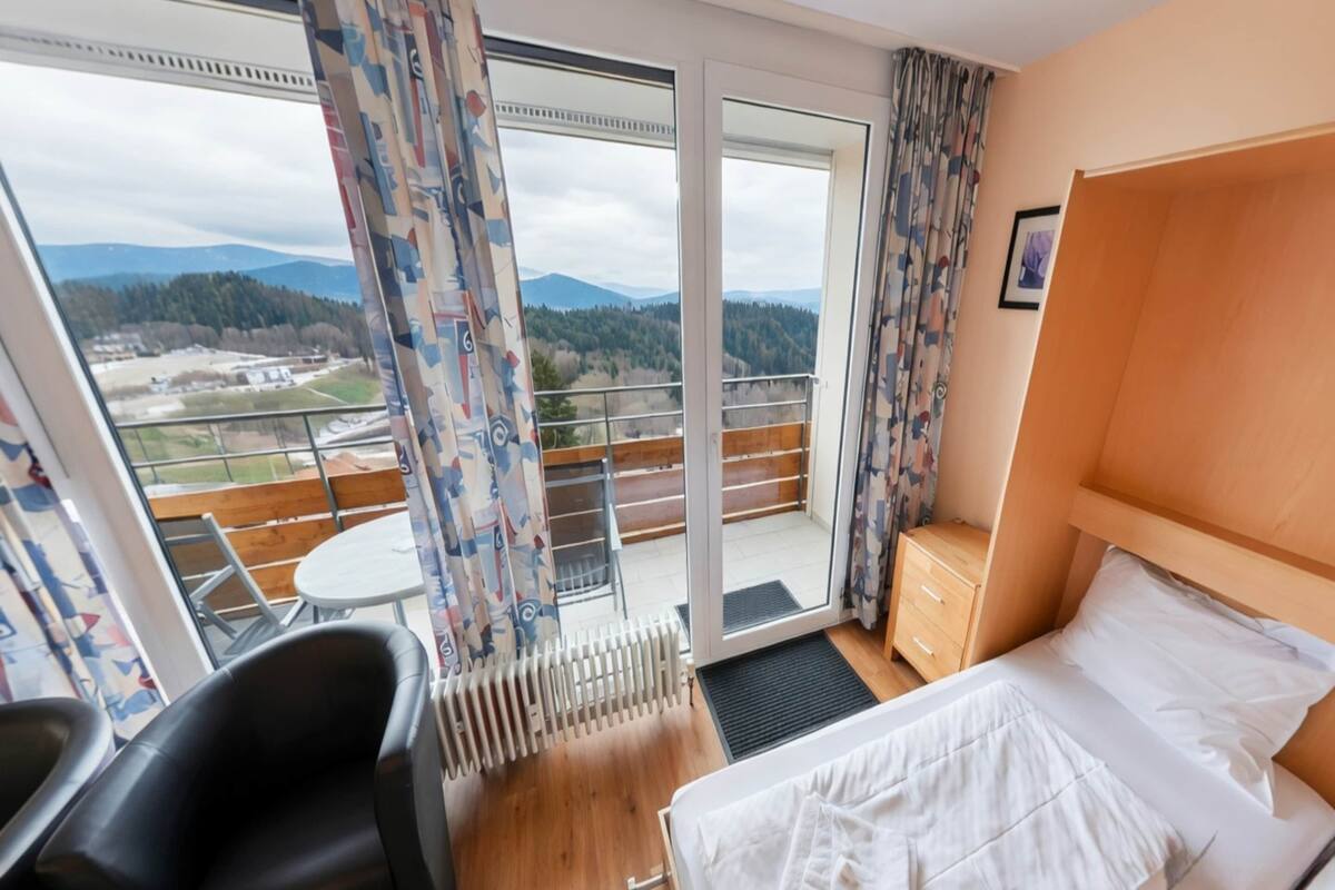 Habitación de hotel con cama, una silla, una mesita pequeña, una mesita de noche de madera y vistas al exterior a través de una puerta corrediza de vidrio.