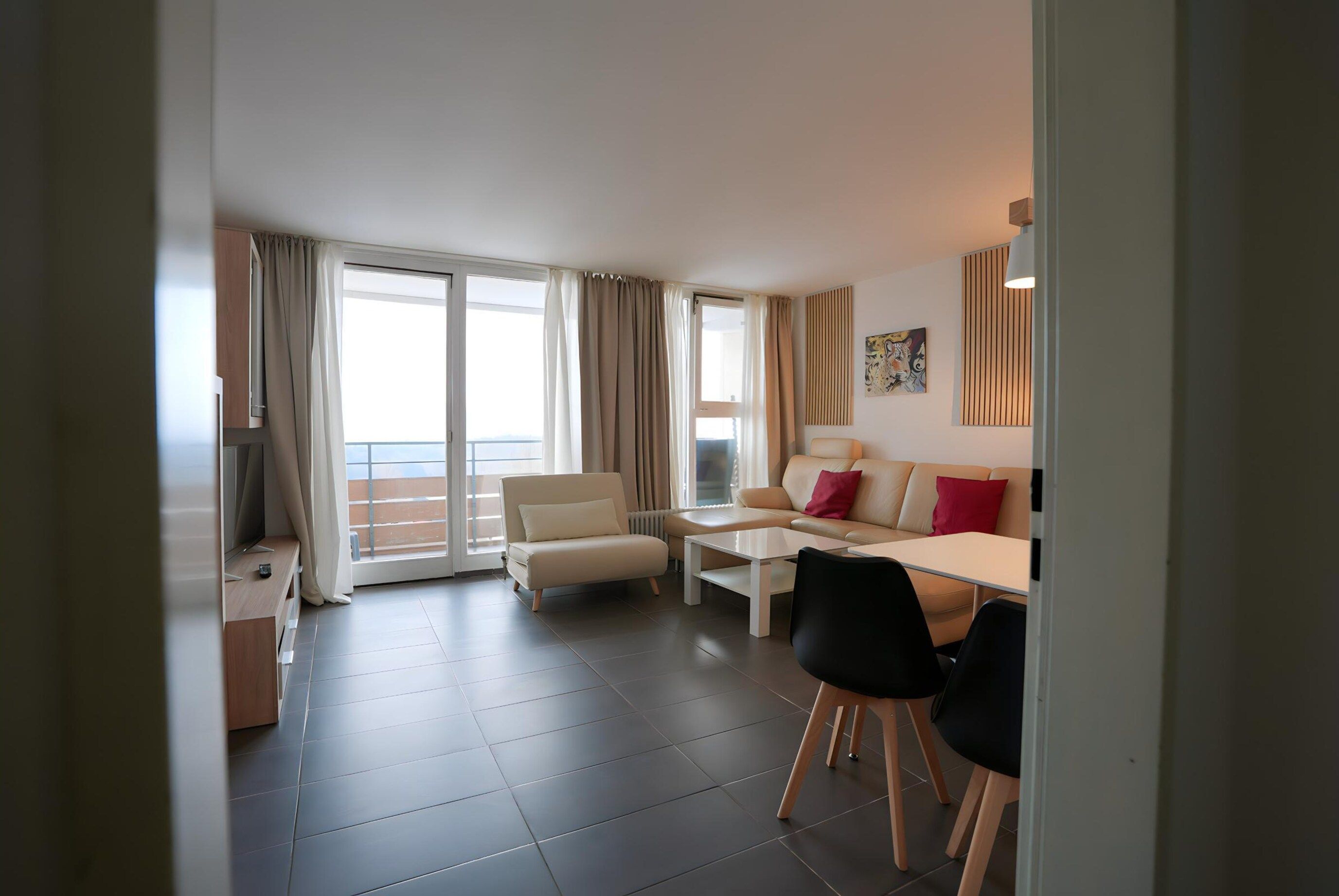 A modern living room with a sofa, coffee table, and chairs, a balcony with curtains, and a large window.