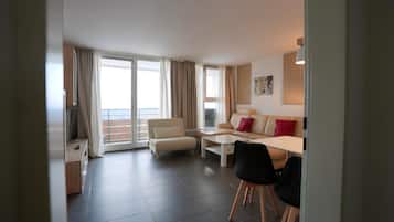 A modern living room with a sofa, coffee table, and chairs, a balcony with curtains, and a large window.