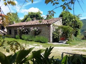 A stone building with a tiled roof, surrounded by greenery and a clear sky.