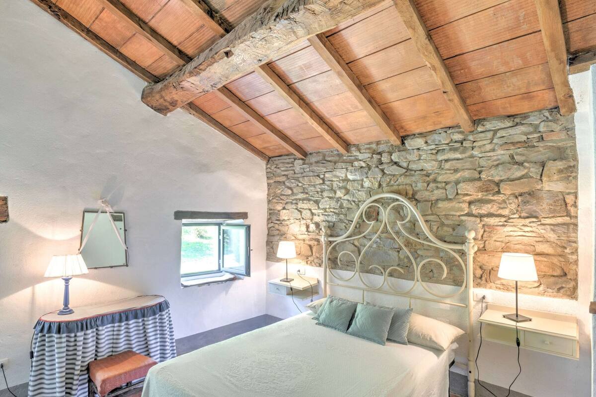 A bedroom with a stone wall, wooden ceiling beams, a bed with pillows, bedside lamps, and a small table with a lamp.