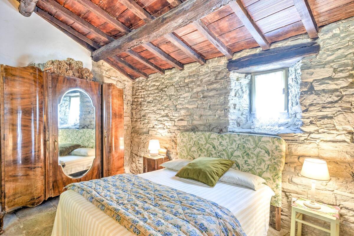 A bedroom with a stone wall, wooden ceiling beams, a bed with floral bedding, a wooden wardrobe, and a small window.