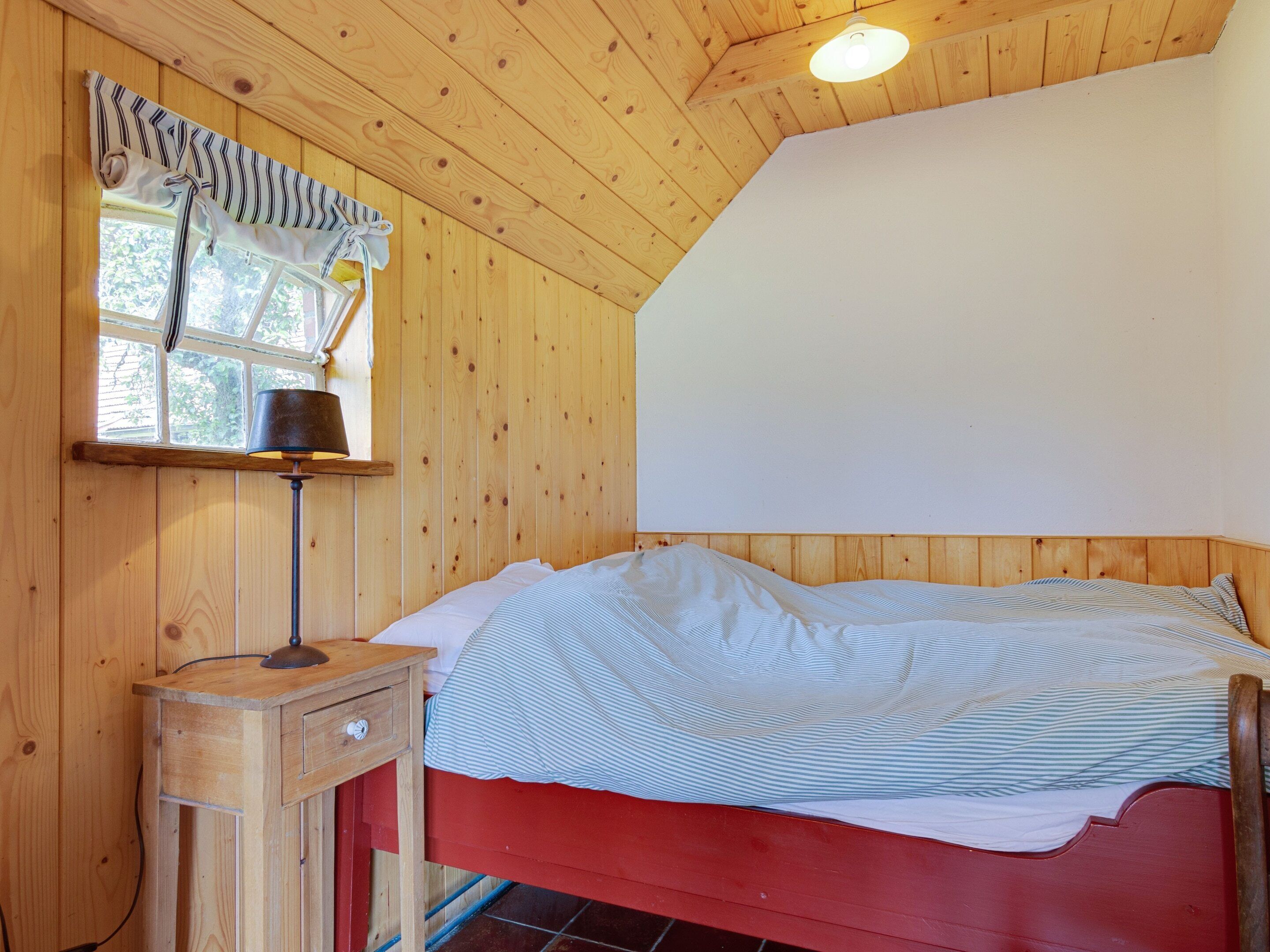 A wooden room with a bed, a nightstand, a lamp, and a window with curtains.