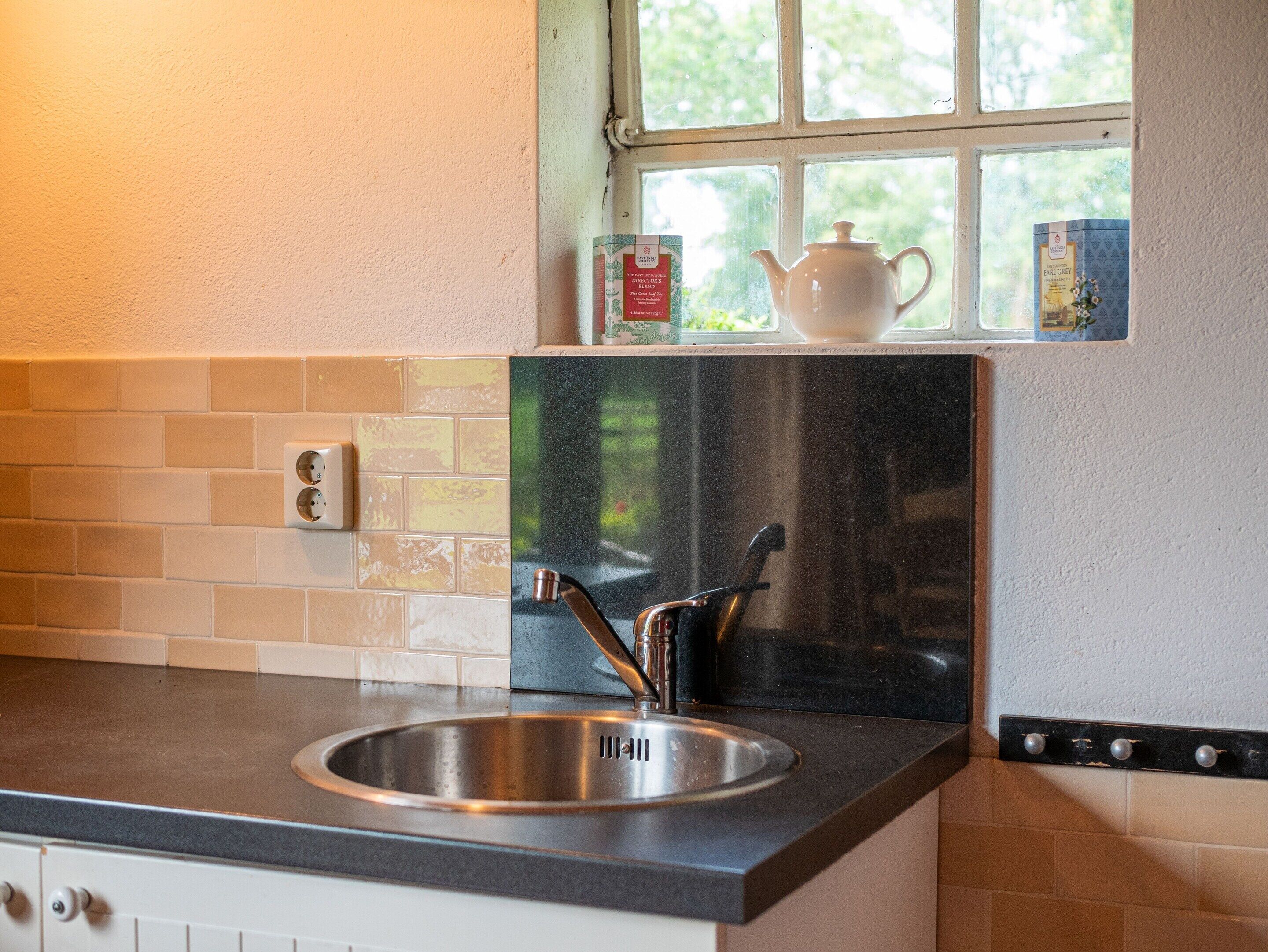 Una cocina con un lavamanos de acero inoxidable, una ventana que da a vistas de follaje, y una tetera blanca en el alféizar.