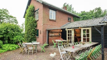 A rustic brick house with a patio area featuring a wooden table and chairs, surrounded by greenery.