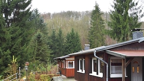 A wooden cabin with a red roof, surrounded by a garden with a paved walkway, and dense forest in the background. - Welcoming Bungalows in Neustadt (Harztor)