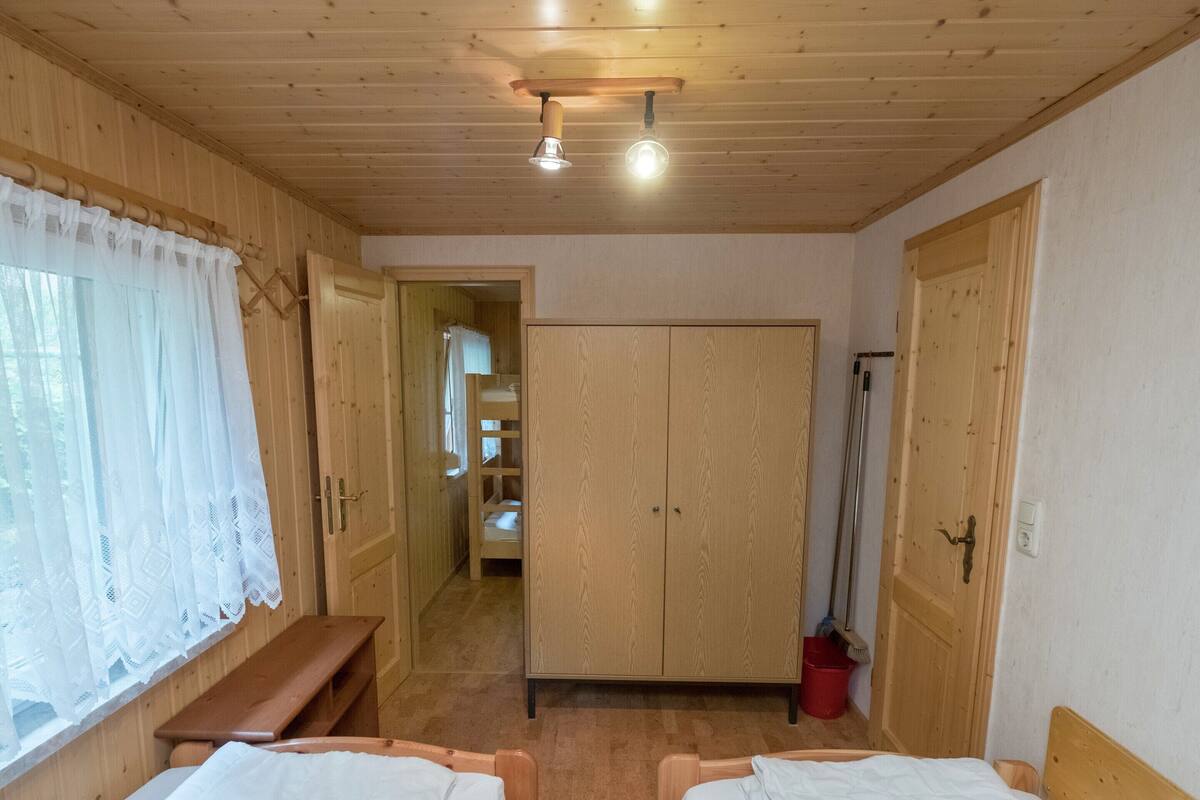 Ein Zimmer mit Holzboden, zwei Betten, einem Kleiderschrank, einer Bank und einem Fenster mit Vorhängen.