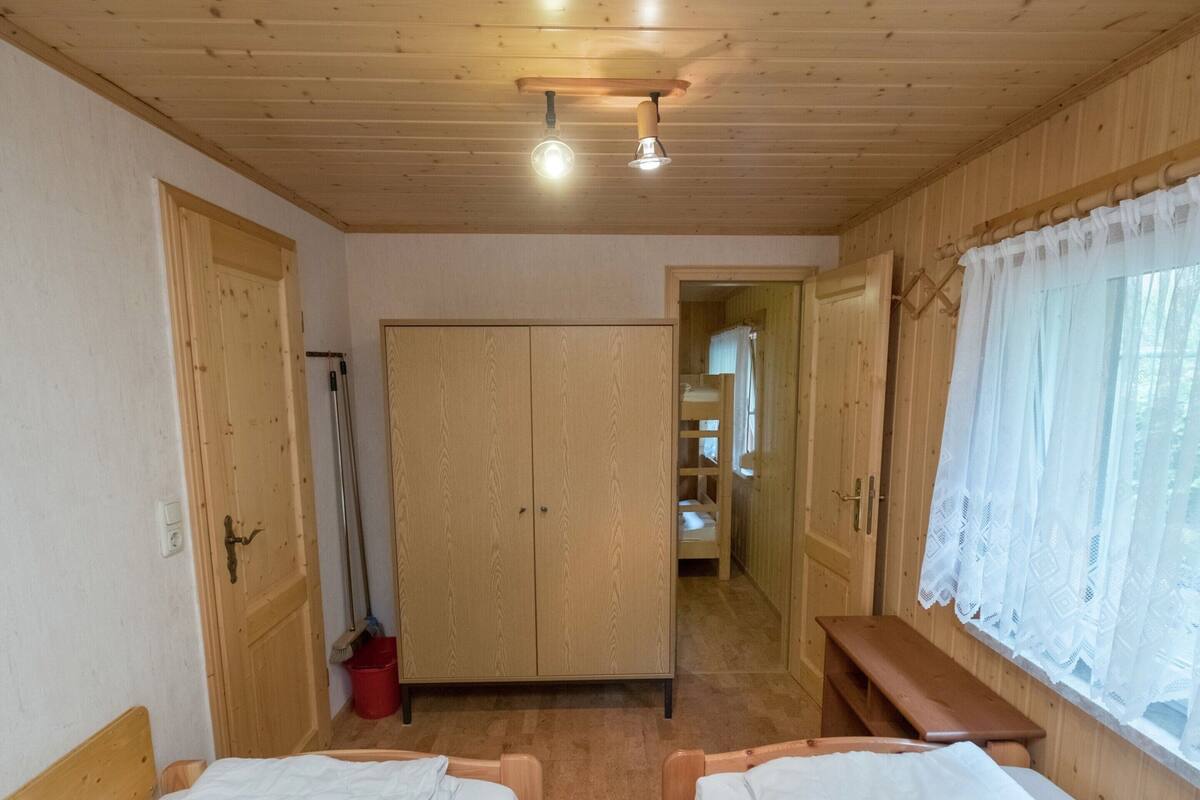Ein Zimmer mit zwei hölzernen Betten, einem Schrank, einer Bank und einem Fenster mit Spitzenvorhängen.