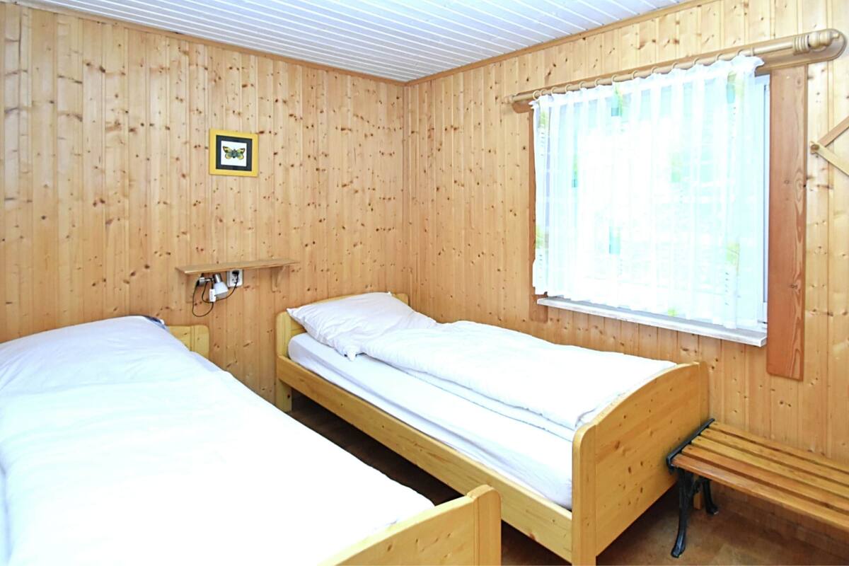 Ein Zimmer mit zwei Holzbettställen, einem kleinen Regal mit Bilderrahmen, einem Fenster mit Vorhängen und einer Holzbank.