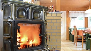 A wood-burning stove with visible flames, a basket of firewood, and a dining area with a table and chairs in the background. - Welcoming Bungalows in Neustadt (Harztor)