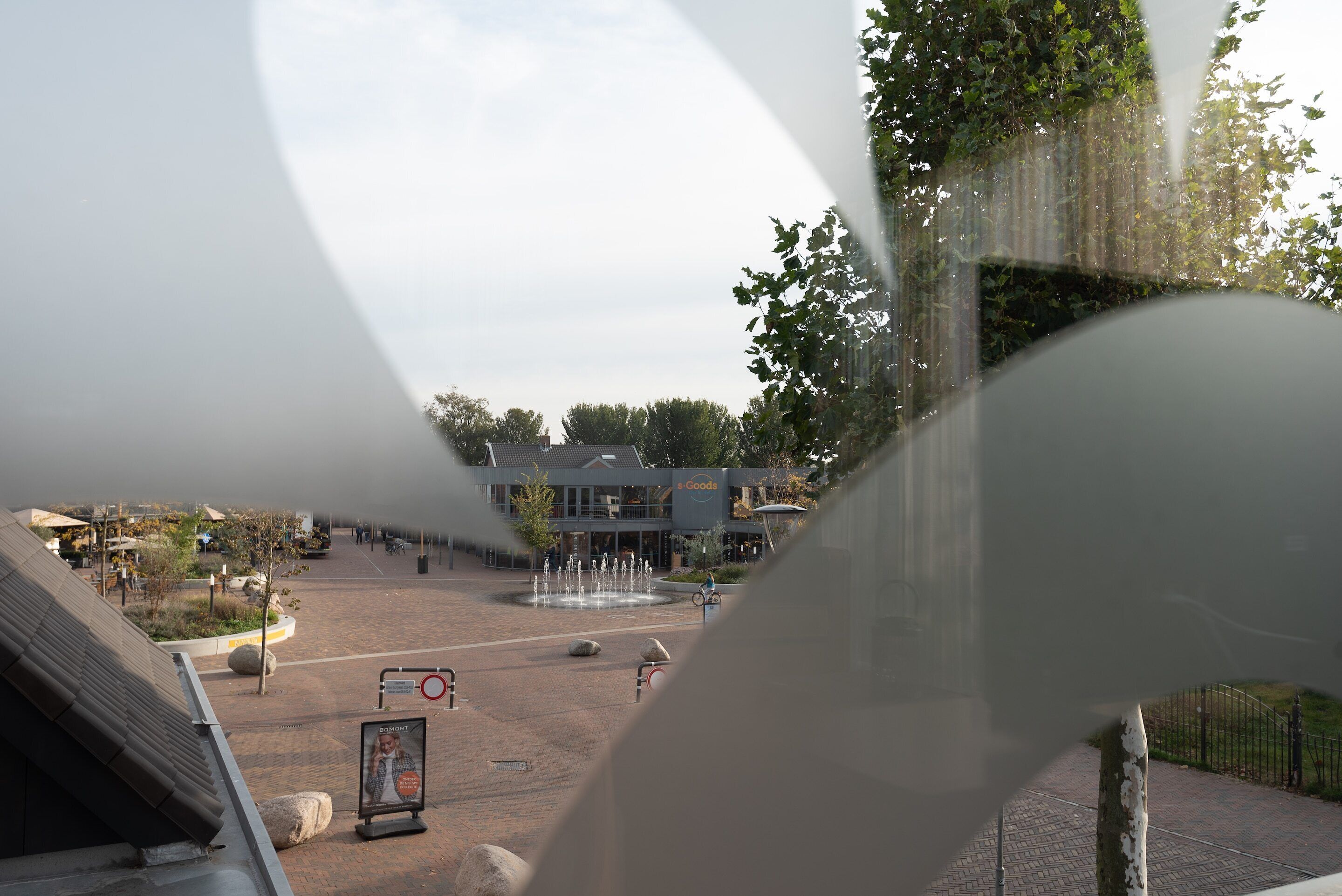 Ett torg med en fontän, en byggnad med skylten ”s Goods”, och en anslagstavla.