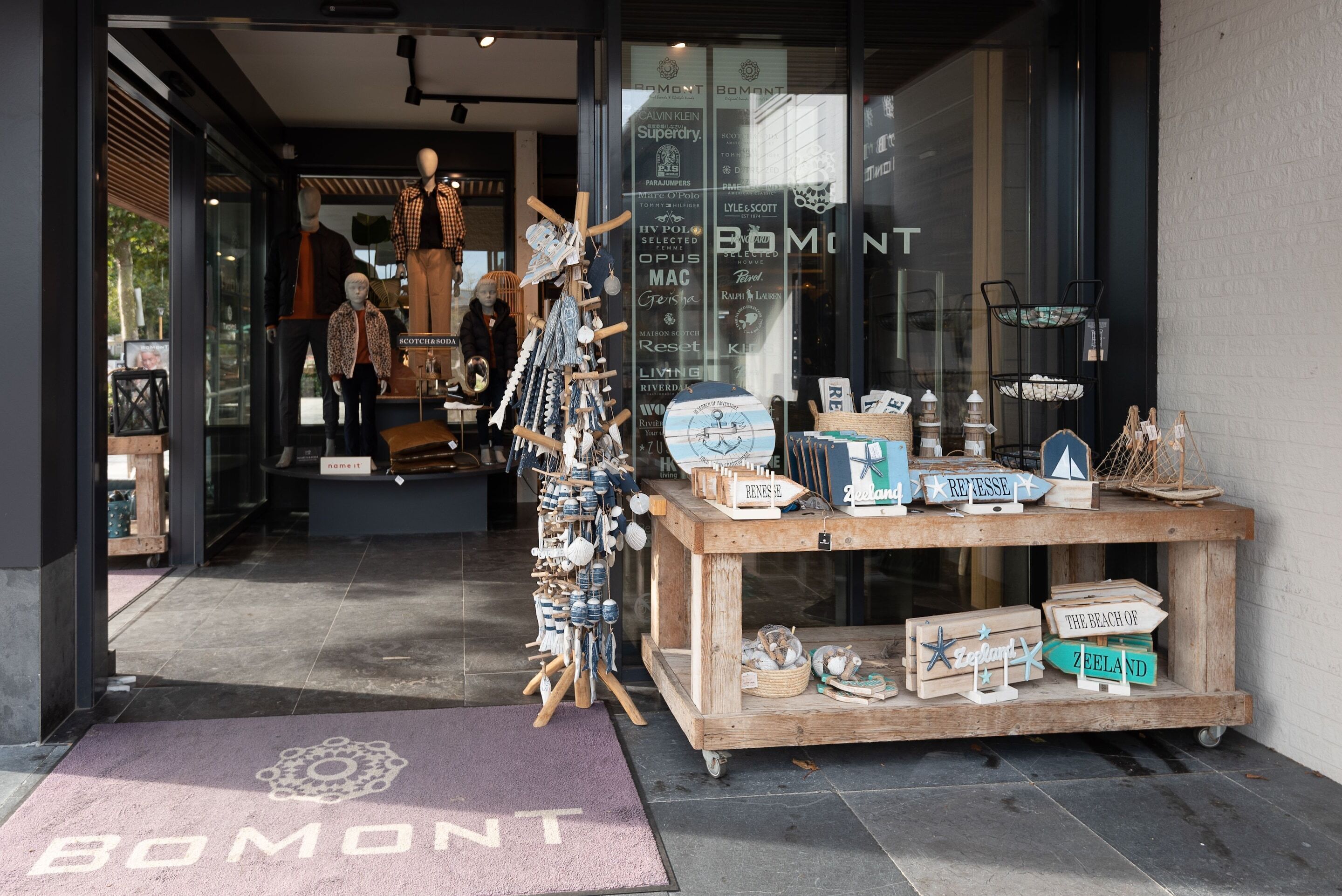 Une vitrine avec des mannequins présentant des vêtements, une table d’exposition en bois sur laquelle sont disposés divers articles, et une enseigne indiquant « BOMONT ».