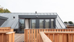 Una terraza de madera con techo metálico y un ventanal corredera que da a una habitación de color oscuro.