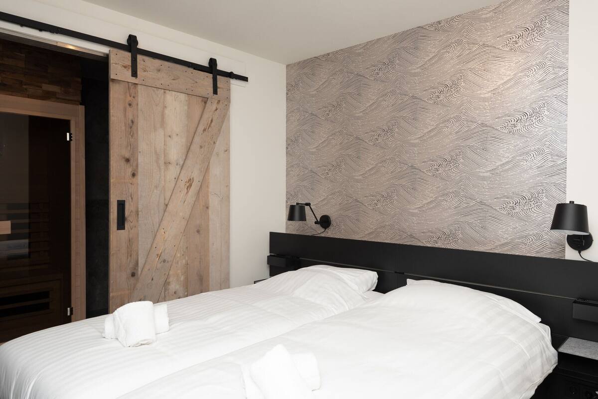 A hotel room with a large bed, a wooden sliding door, a patterned wall, and a bedside table with a lamp.