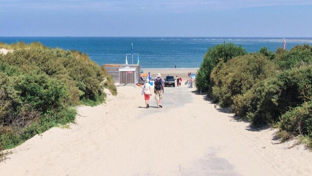 Un sendero arenoso que conduce a una playa, con una pequeña construcción, probablemente un baño, e izada una bandera.