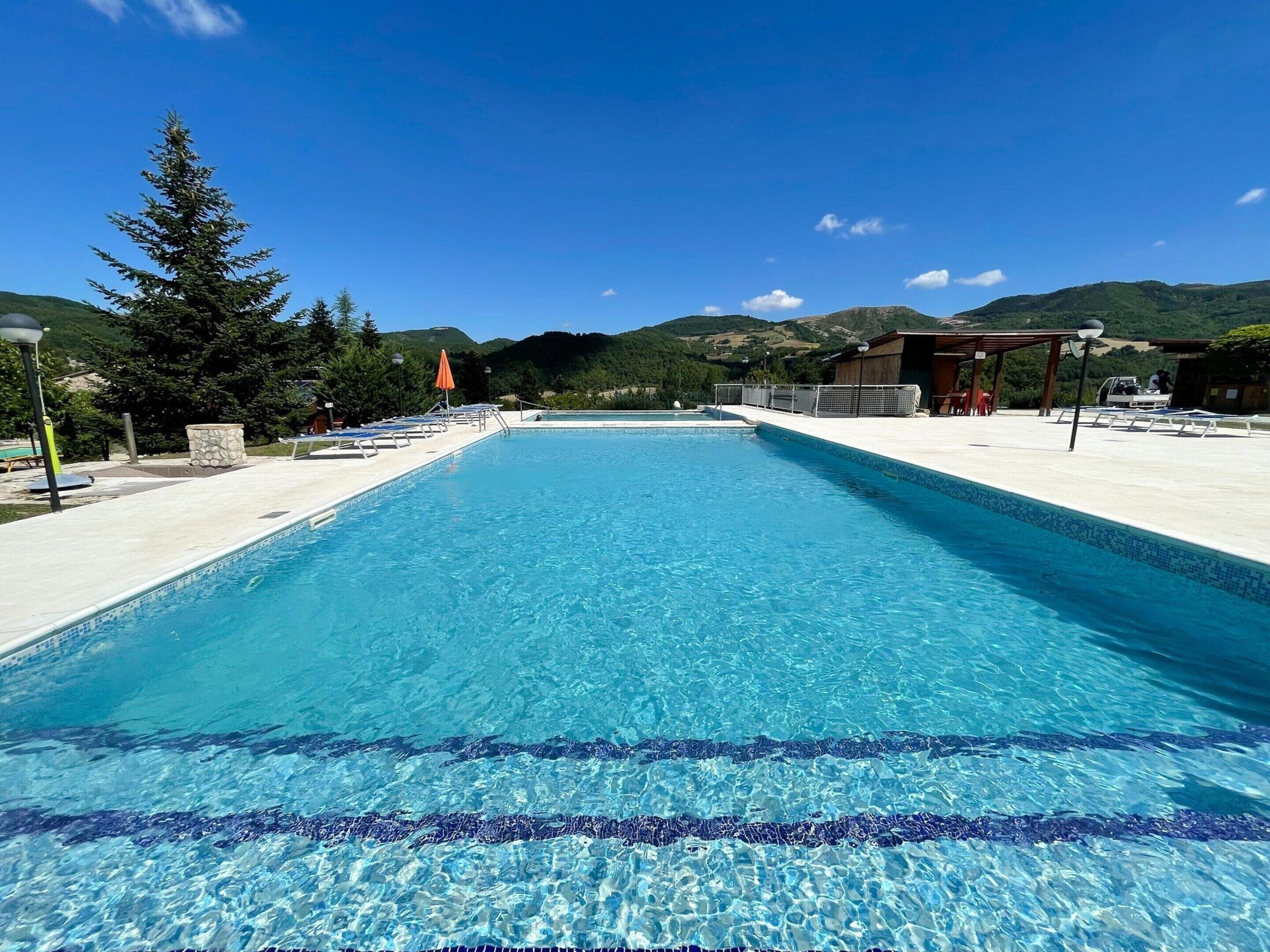 Ein rechteckiger Pool mit klarem, blauem Wasser, umgeben von einer weißen Liegefläche. Auf der Liegefläche stehen Liegestühle und Sonnenschirme, und im Hintergrund ist eine hölzerne Konstruktion mit Dach zu sehen.