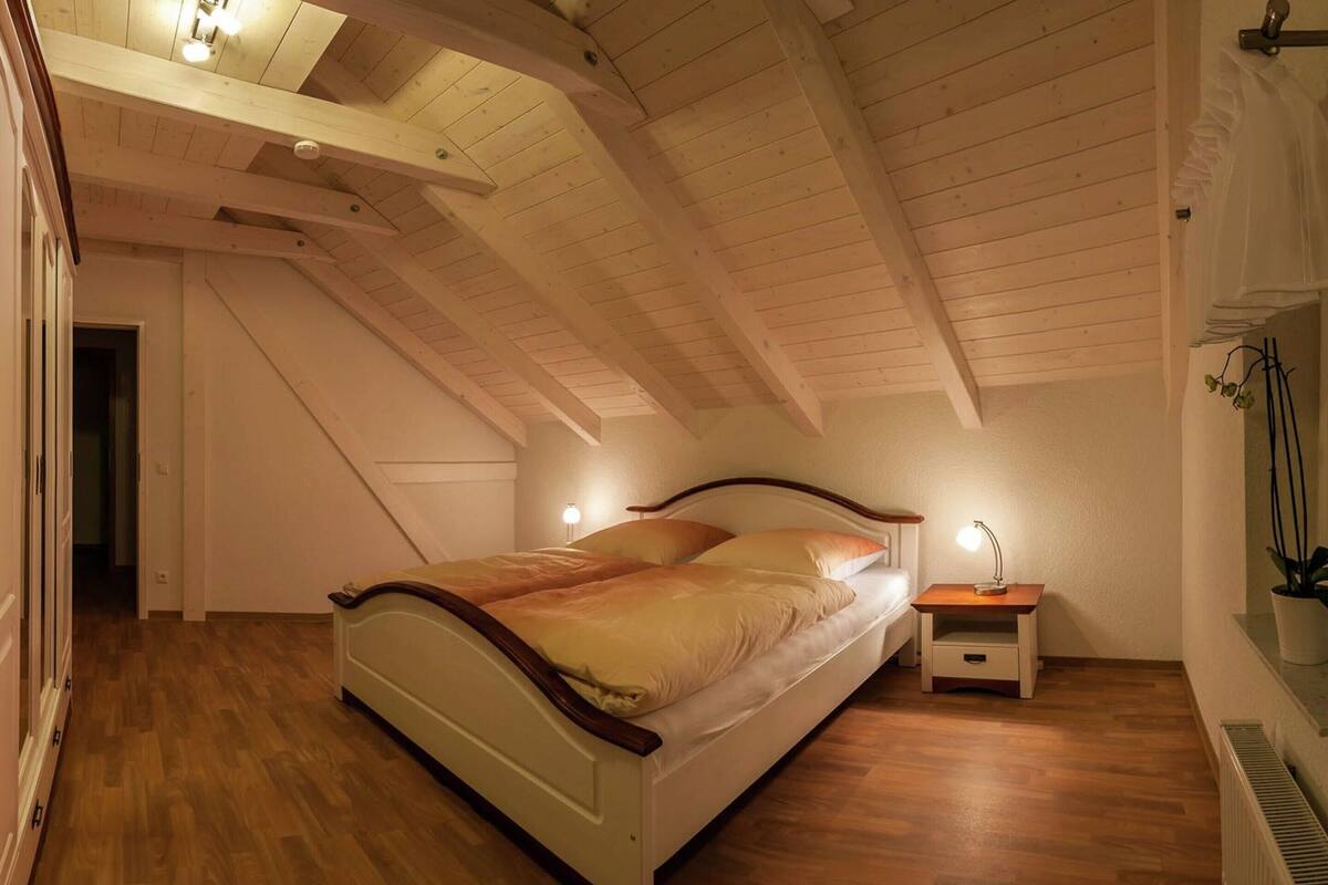 A bedroom with a sloped ceiling, wooden floor, a bed with white bedding, a bedside table with a lamp, and a potted plant.