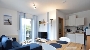 Ein modernes Wohnzimmer mit einer Couch, einem Esstisch mit Stühlen, einem Fernseher und einem Balkon mit Vorhängen.