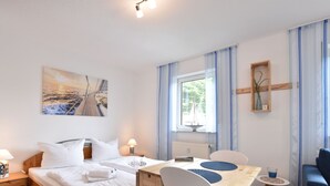 Ein Hotelzimmer mit einem Bett, einem hölzernen Schreibtisch, zwei Stühlen, einem kleinen Tisch, einem Fenster mit Vorhängen, einer Wanduhr und einem Gemälde.