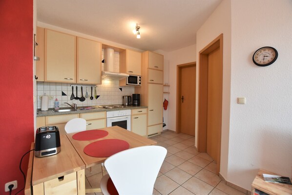 A compact kitchen with a toaster, microwave, and sink. There is a wooden table with two chairs and a red placemat.