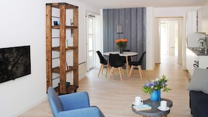 A modern living room with a blue armchair, a coffee table with a book and a cup, a wooden bookshelf, a dining area with a table and chairs, and a kitchen in the background. - 4 Person Holiday Home in Allinge (Allinge)