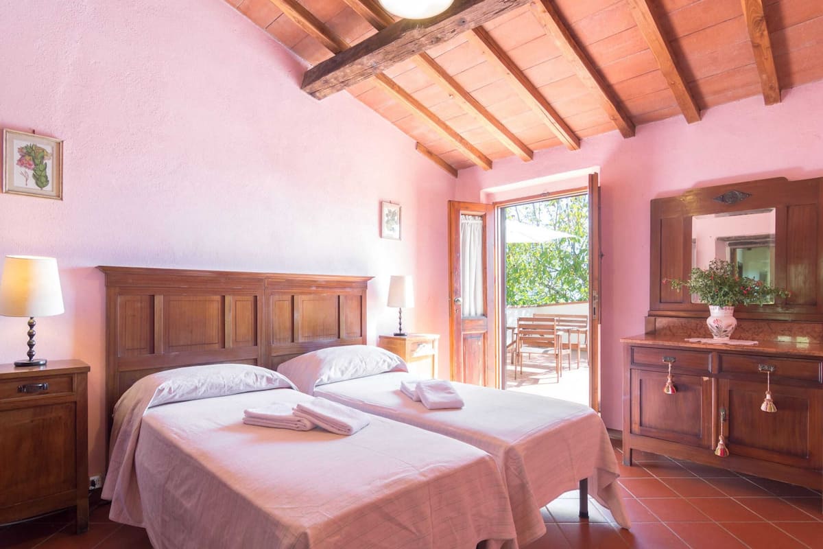 A bedroom with two beds, a wooden headboard, a nightstand with a lamp, a dresser with a mirror, and a balcony with outdoor seating.