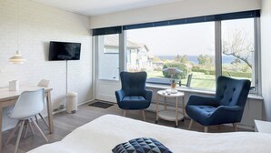 A modern hotel room with a large bed, two blue armchairs, a desk with a chair, a TV mounted on the wall, and a view of the sea through large windows. - 2 Person Holiday Home in Allinge (Allinge)