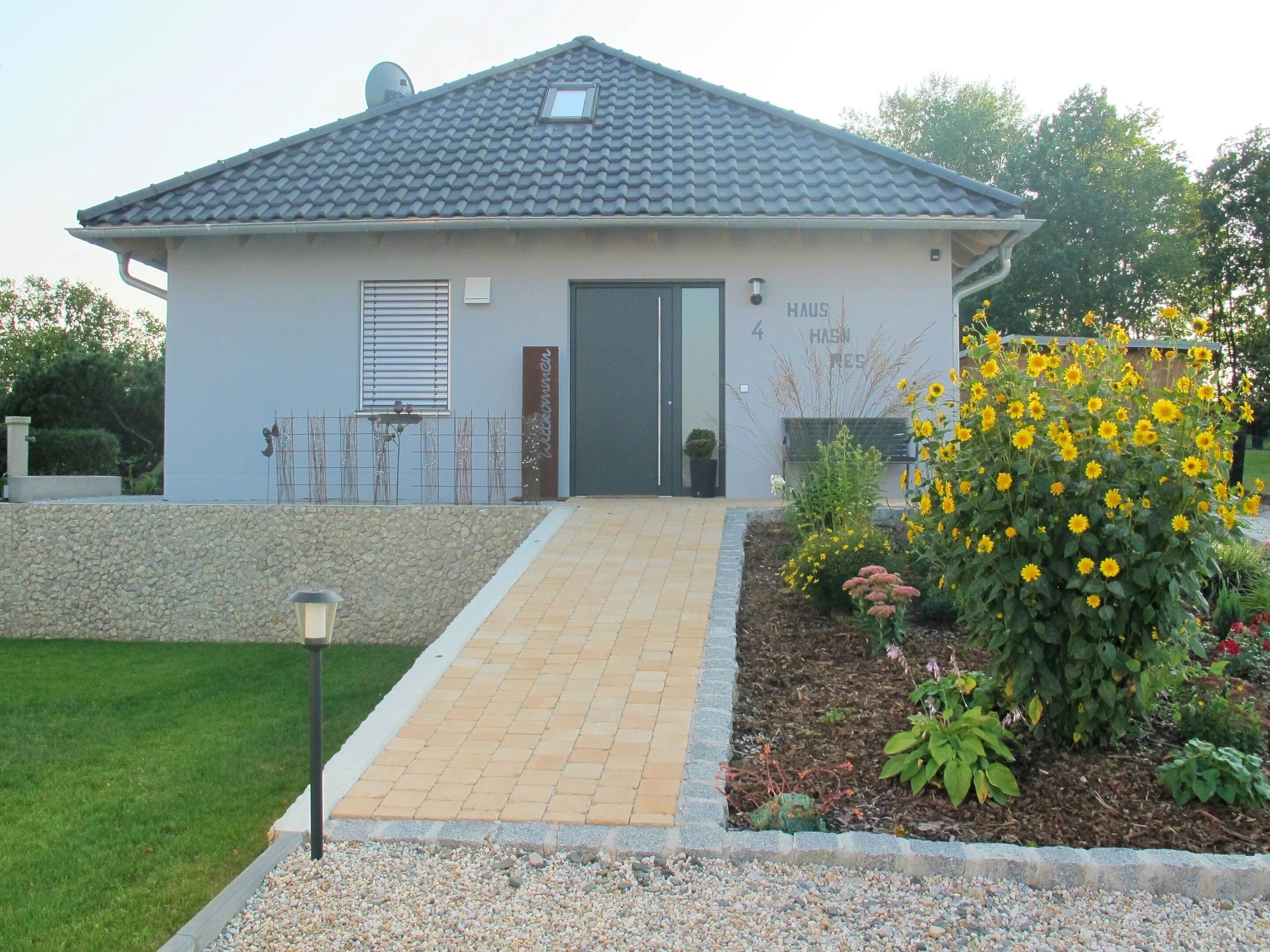 A single-story building with a tiled roof, a front door, and a small garden with yellow flowers.