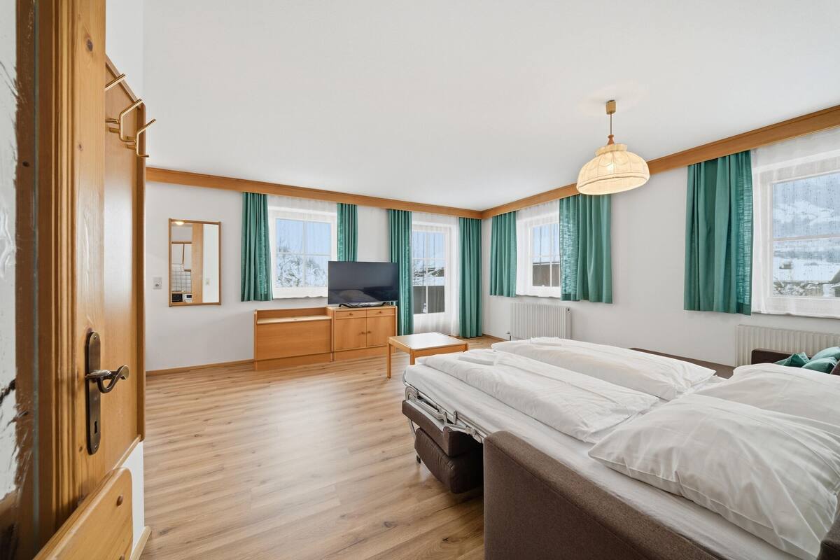 A hotel room with a wooden floor, a bed with white linens, a TV on a wooden desk, and large windows with green curtains.