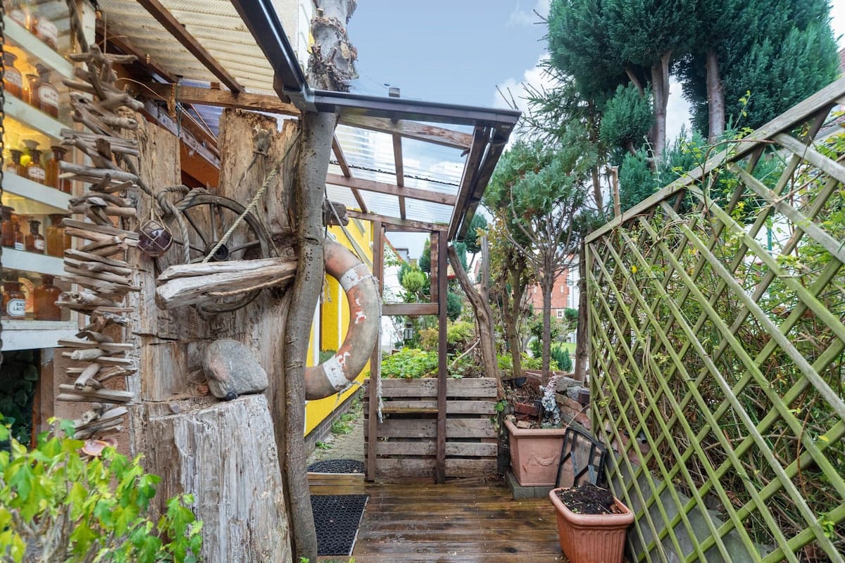 A narrow wooden pathway leading to a wooden structure with a yellow door, surrounded by potted plants and a wooden fence.