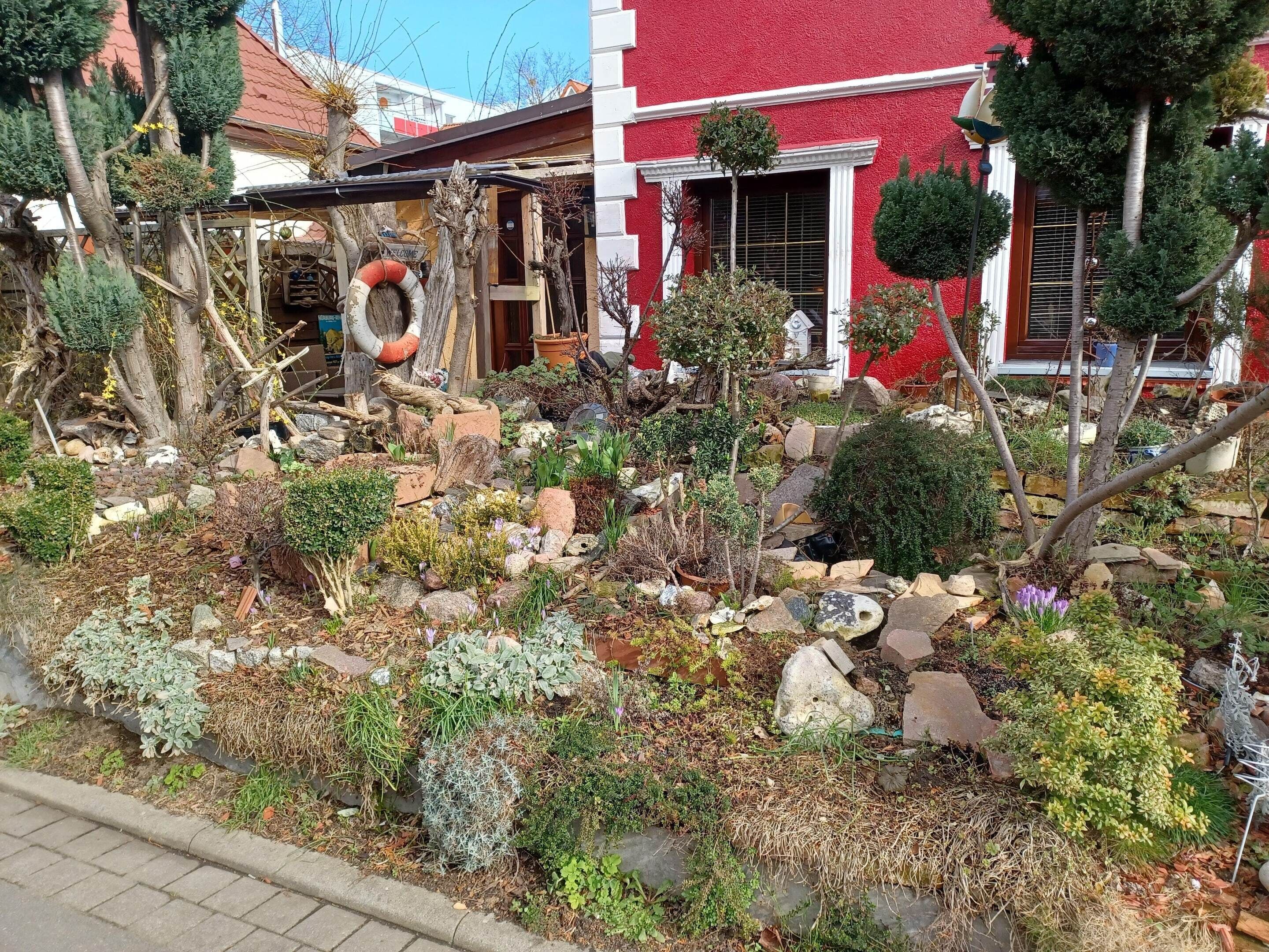 Un jardin avec différentes plantes et des rochers, une structure en bois et, au fond, un bâtiment rouge.