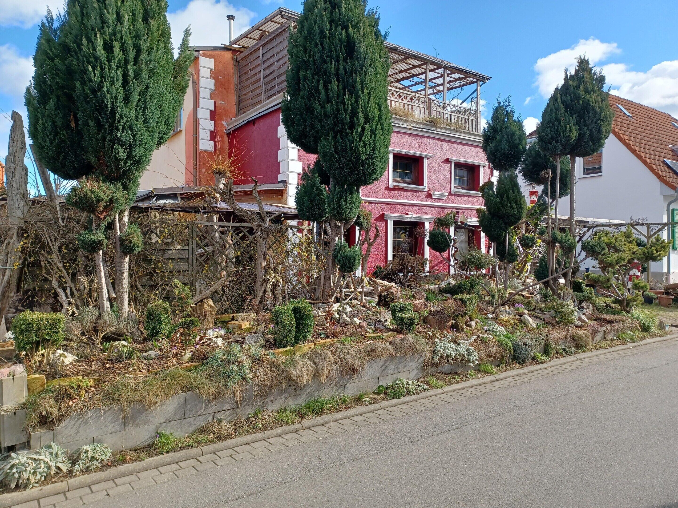 A residential building with a red facade, a wooden pergola on the roof, and a garden with various plants and trees.