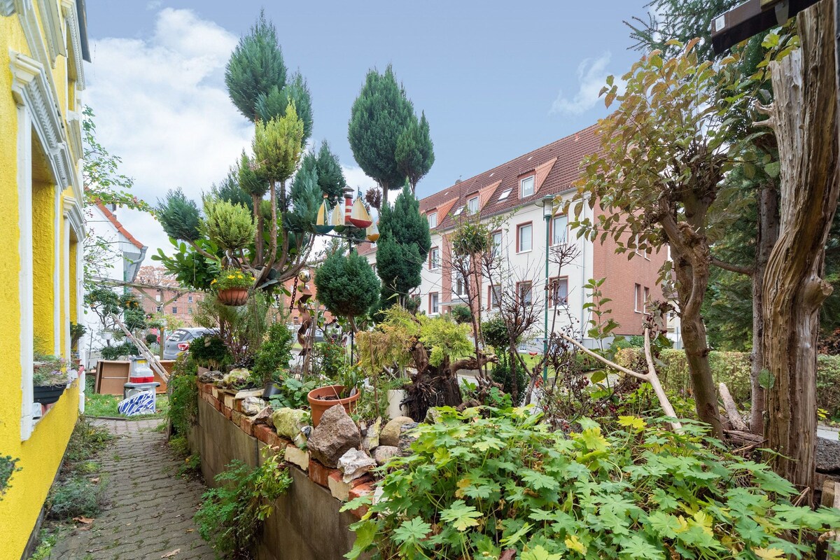A narrow alleyway with a yellow building on the left, a stone wall with plants, and a row of trees and shrubs.
