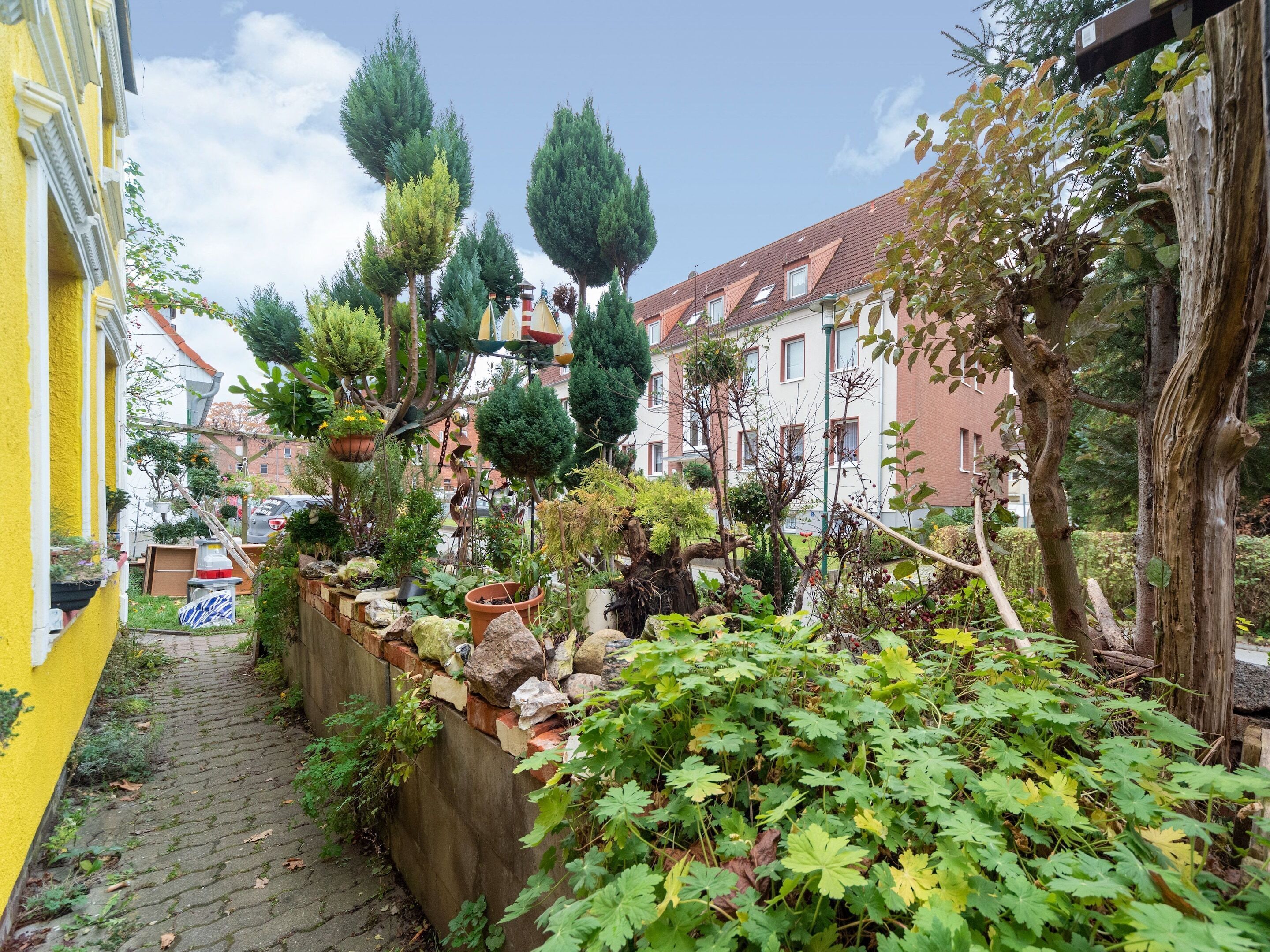 Ein schmaler Gang mit einem gelben Gebäude auf der linken Seite, einer Steinmauer mit Pflanzen und einer Reihe Bäume und Sträucher.