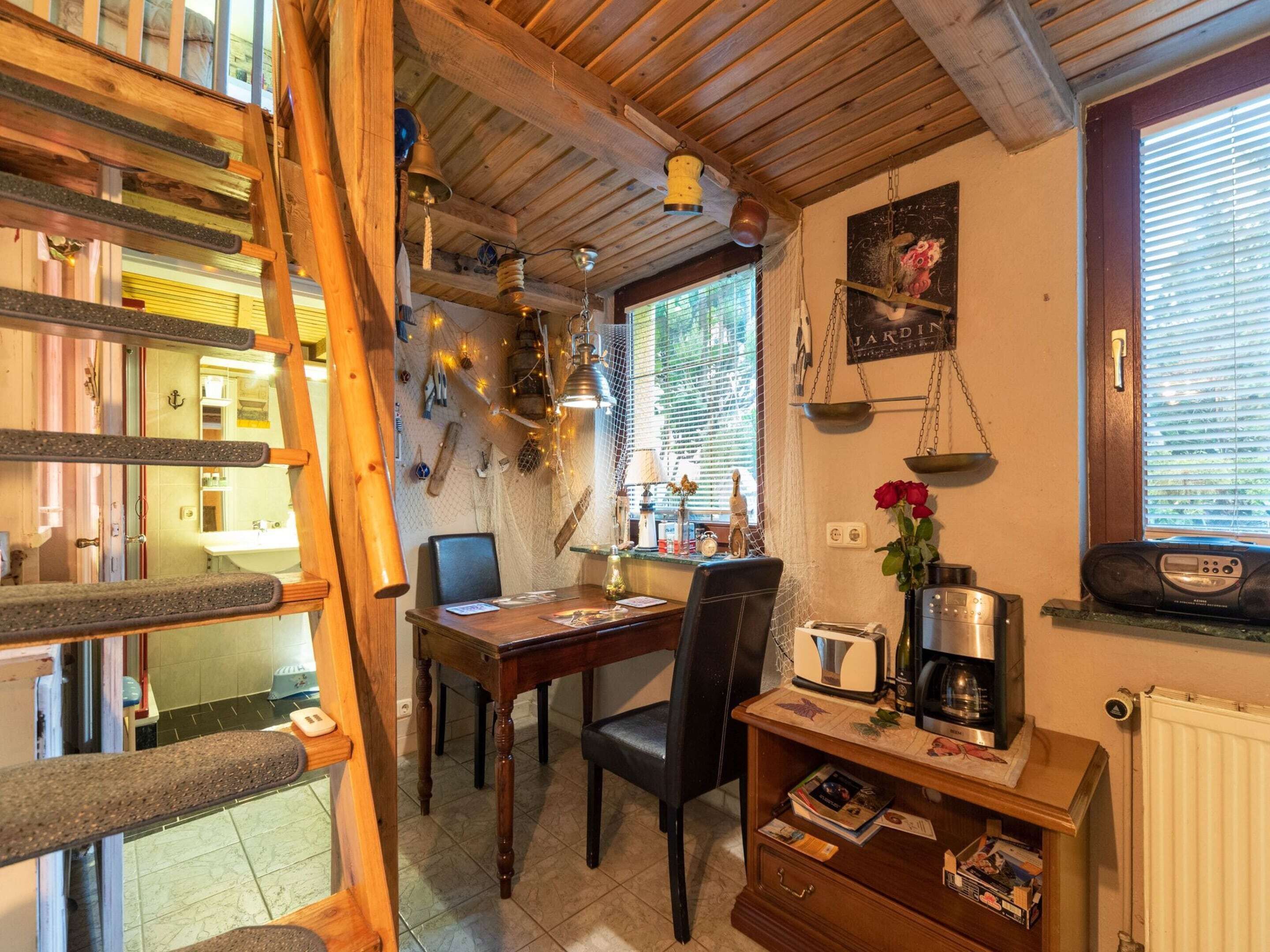 A cozy kitchen with wooden beams, a dining table, chairs, a coffee machine, and a small shelf with books and a vase of flowers.