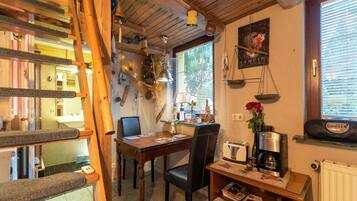 A cozy kitchen with wooden beams, a dining table, chairs, a coffee machine, and a small shelf with books and a vase of flowers.