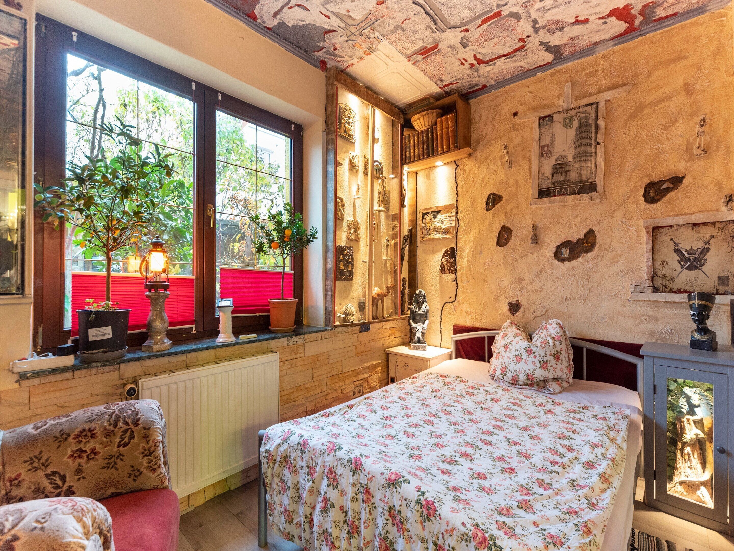 A room with a bed, a window with plants, a radiator, and a wall with various decorations.